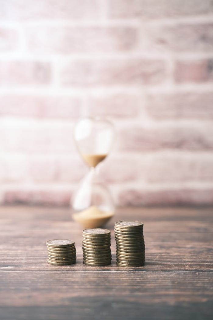 Stacks of coins with an hourglass in the background symbolize savings and time management.